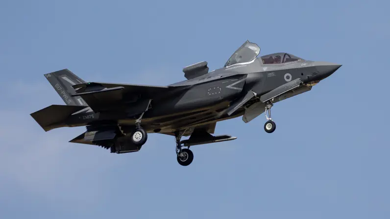  Een zwarte straaljager met open cockpit en ingetrokken wielen, vliegend tegen een heldere blauwe lucht met zichtbare markeringen en nummers.    