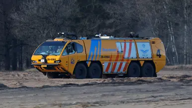 Een grote gele brandweerauto met zes wielen, blauwe en rode strepen, rijdt over een zanderig terrein in een bos. 