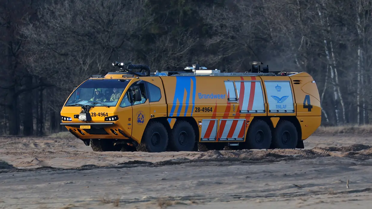 Een grote gele brandweerauto met zes wielen, blauwe en rode strepen, rijdt over een zanderig terrein in een bos. 