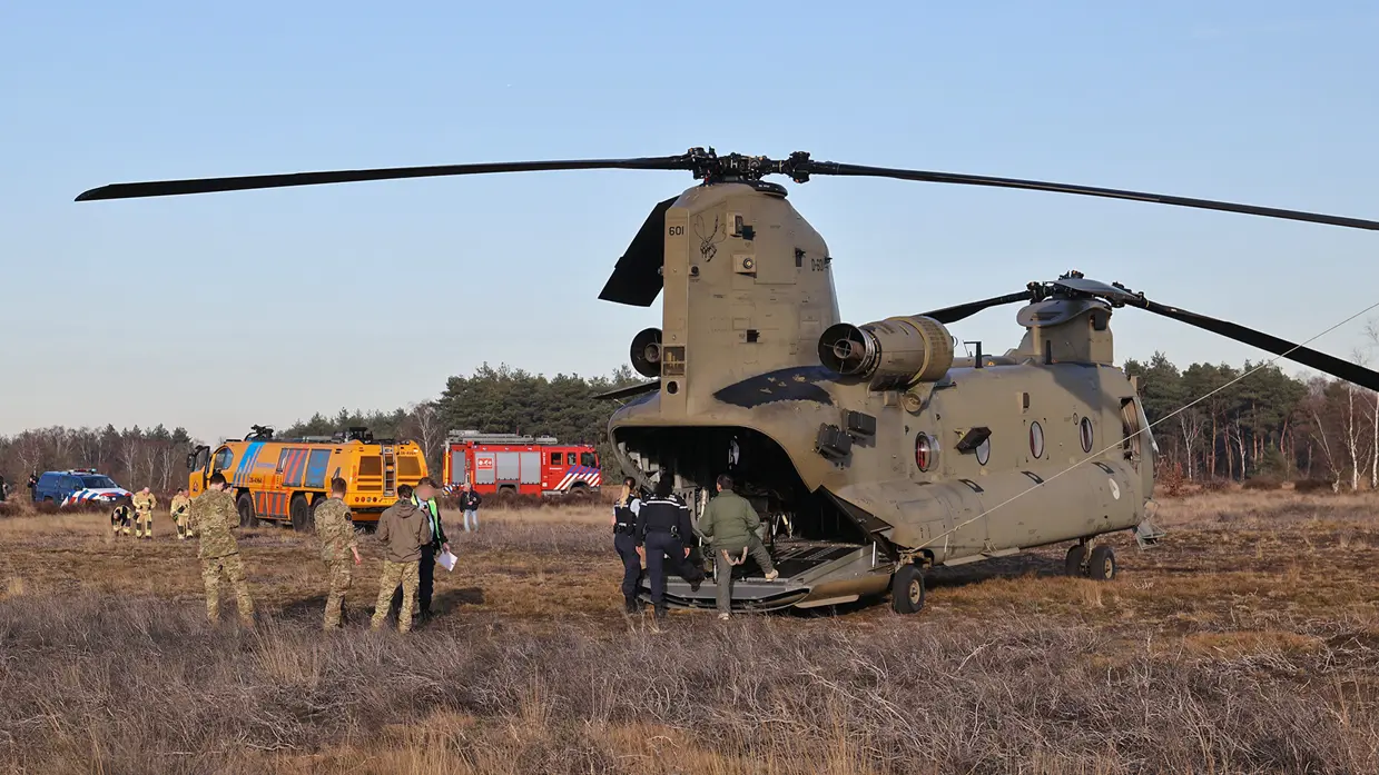 Een grote militaire helikopter met open laaddeur staat op een veld, omringd door voertuigen en mensen in uniform.