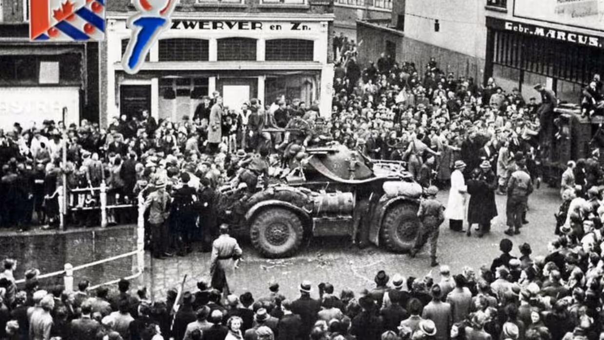 Een grote menigte omringt een militaire pantserwagen op een straat, met gebouwen en een bord met de Canadese vlag op de achtergrond. De foto lijkt historisch.