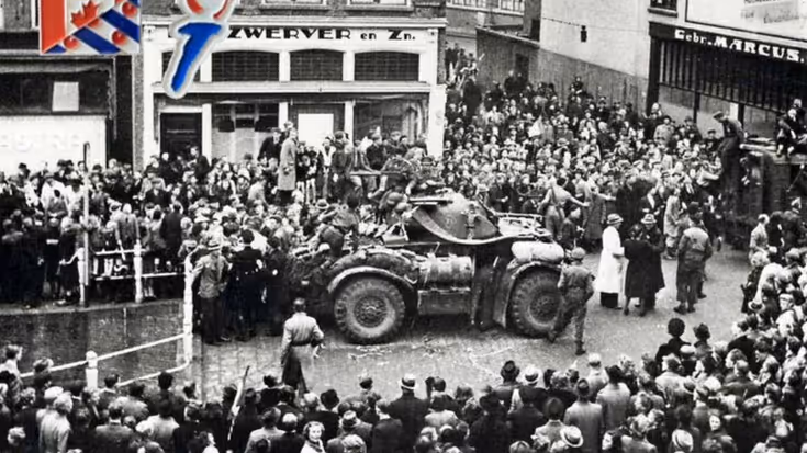 Een grote menigte omringt een militaire pantserwagen op een straat, met gebouwen en een bord met de Canadese vlag op de achtergrond. De foto lijkt historisch.