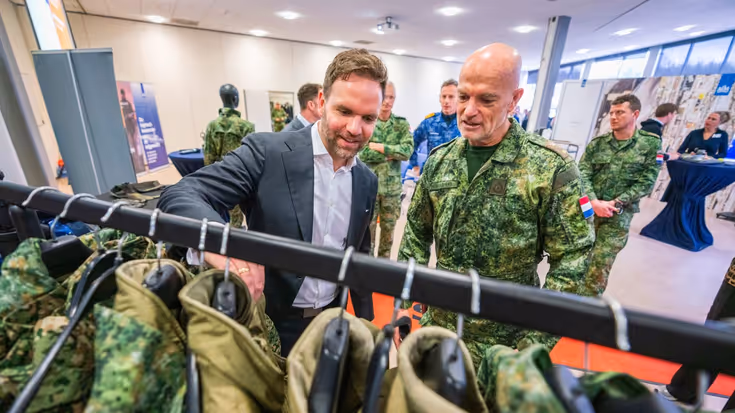 Staatssecretaris Derk Boswijk, in pak, gaat met zijn hand door een kledingrek met militaire uniformen. Een militair kijkt met hem mee.