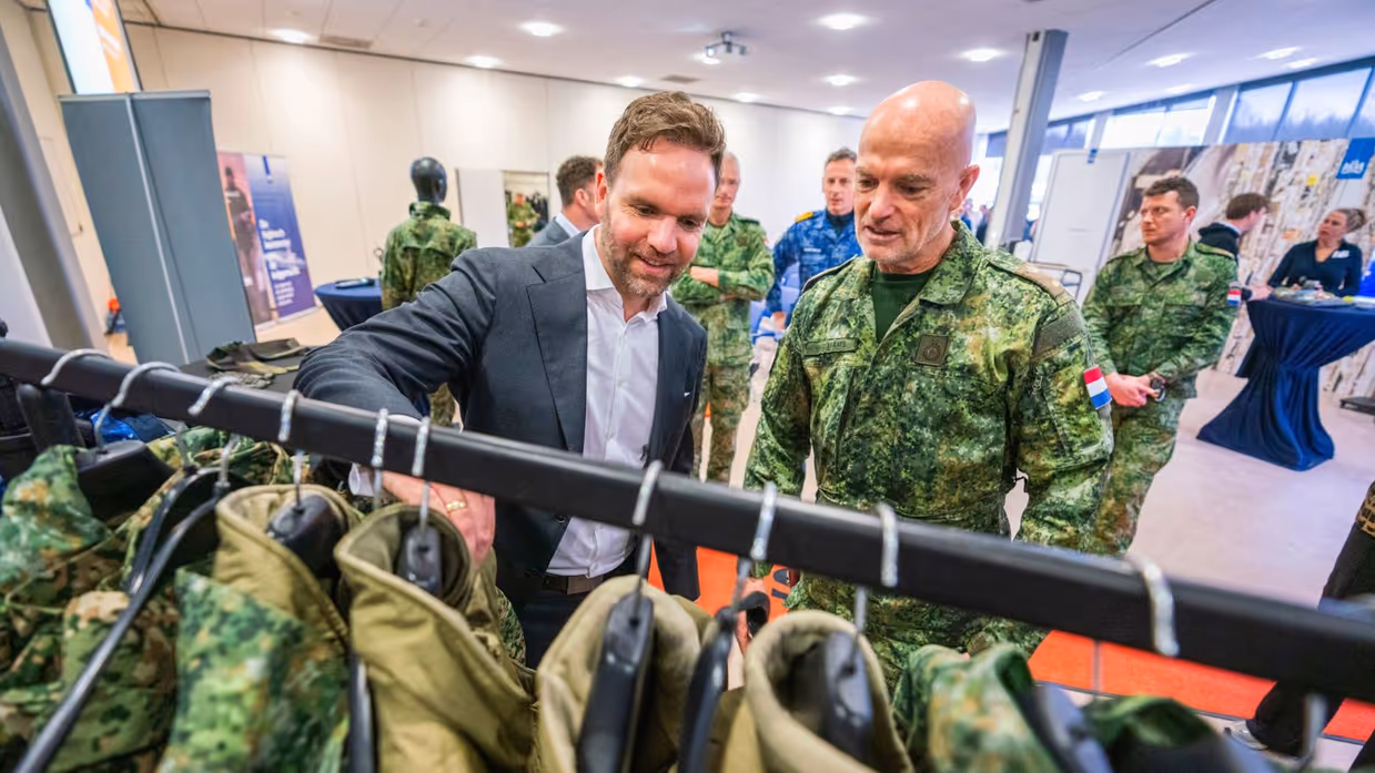 Staatssecretaris Derk Boswijk, in pak, gaat met zijn hand door een kledingrek met militaire uniformen. Een militair kijkt met hem mee.