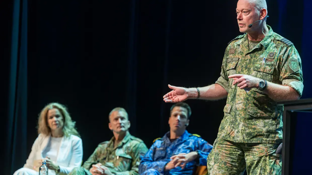 Op het podium staat een commandant die zijn ervaring uit de praktijk deelt met het publiek. Naast hem zitten collega sprekers op het podium.