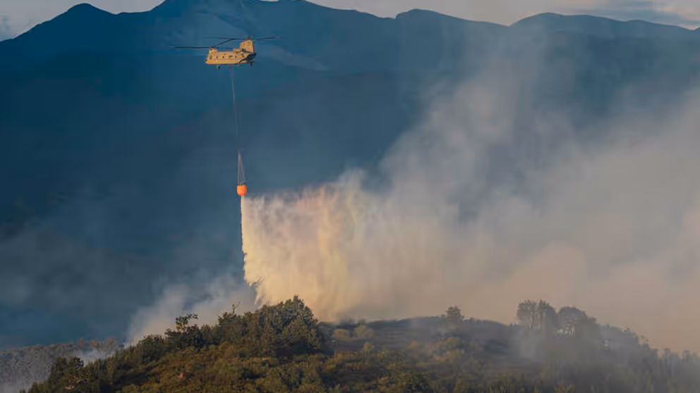 Chinook-helikopter die water uit een oranje bambi bucket laat vallen op brandend terrein.