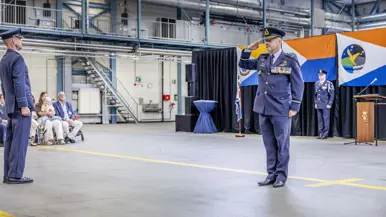 In een ceremonieel aangeklede hangaar brengt Schipmölder de eregroet. Aan de rand van de vloer zit publiek.