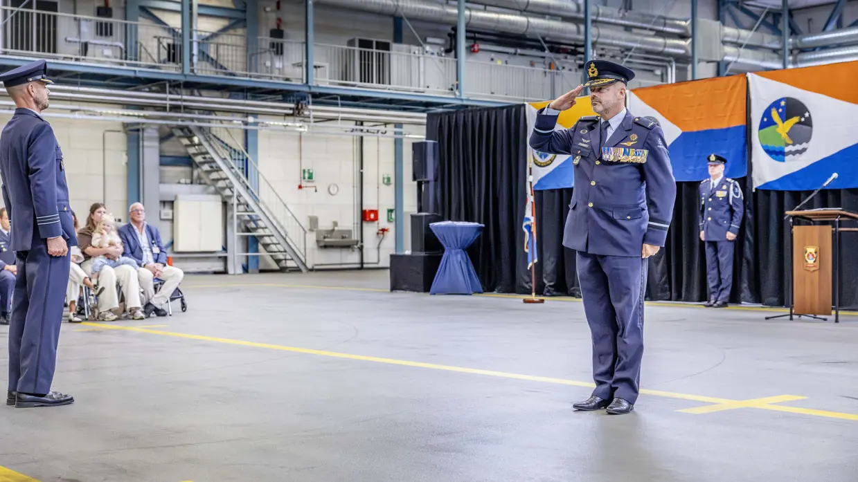 In een ceremonieel aangeklede hangaar brengt Schipmölder de eregroet. Aan de rand van de vloer zit publiek.