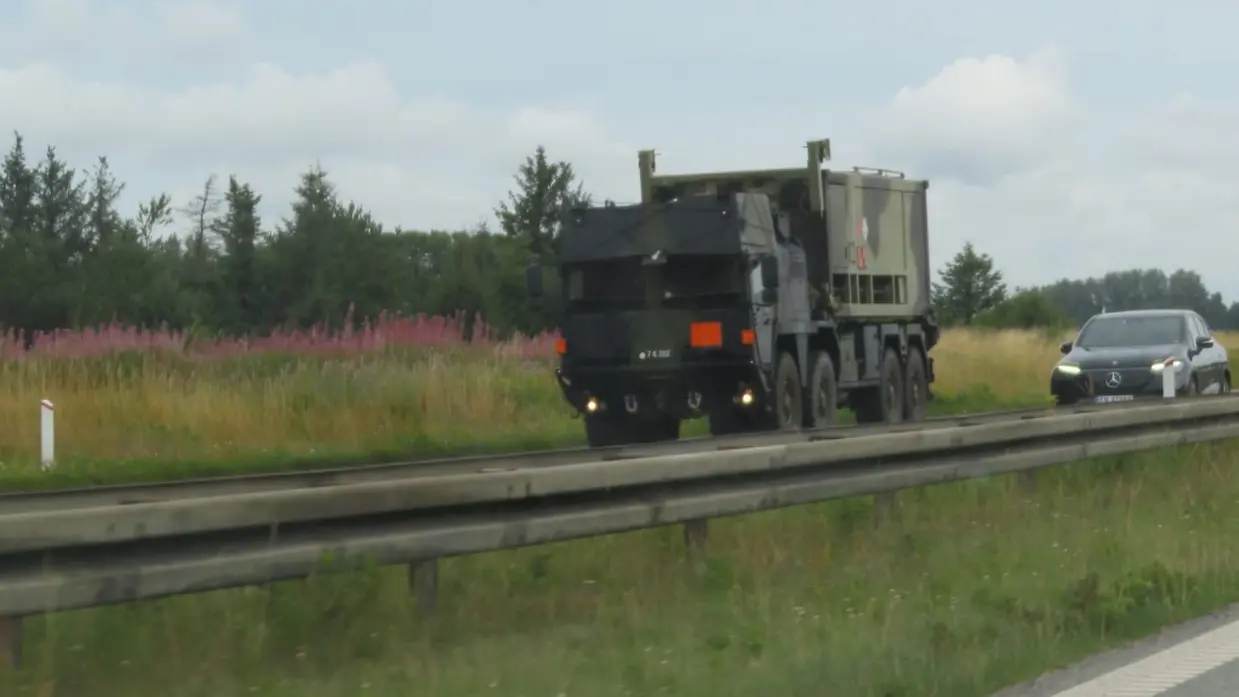 Een grote militaire vrachtwagen rijdt over een weg, omringd door groen gras en paarse bloemen, met een auto erachter.