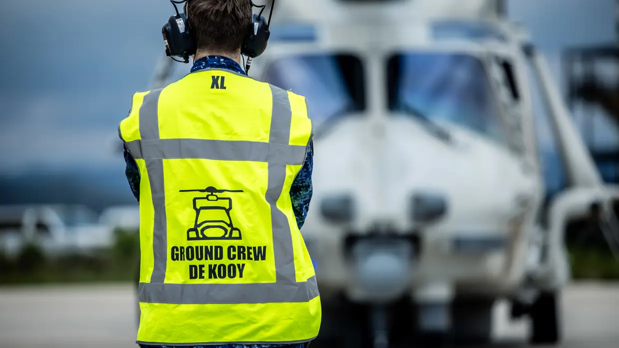 Militair in blauw vlekkenpak met geel veiligheidshesje met op de rug de tekst ‘Ground Crew De Kooy’. Boven de tekst staat een eenvoudige tekening van een vooraanzicht van een NH90. De militair kijkt naar een NH90, die onscherp op de foto staat.