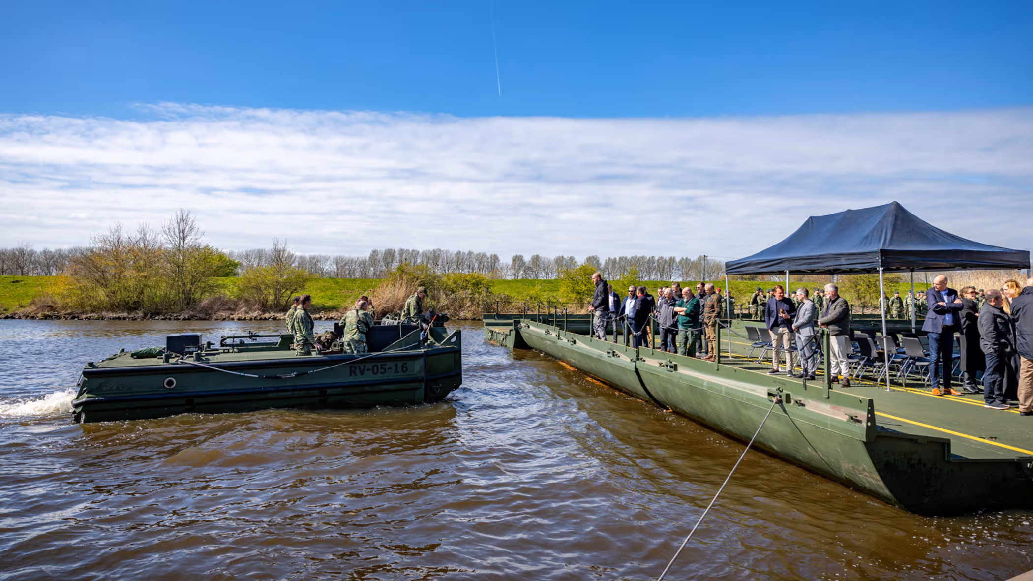 Een groepje militairen op een duwboot vaart richting een legergroen ponton. Daarop staan toeschouwers. Daarboven een zonnige lucht.