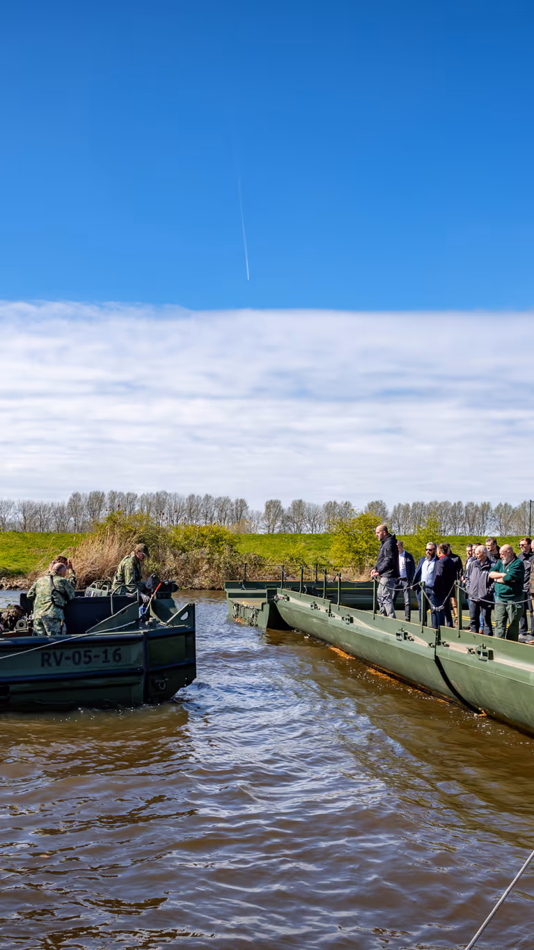 Een groepje militairen op een duwboot vaart richting een legergroen ponton. Daarop staan toeschouwers. Daarboven een zonnige lucht.