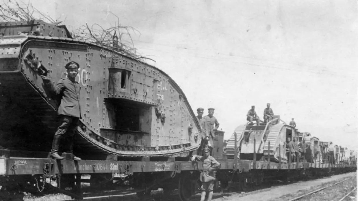 Een zwart-witfoto van een oude tank op een treinwagon, met soldaten erop en ernaast staand.