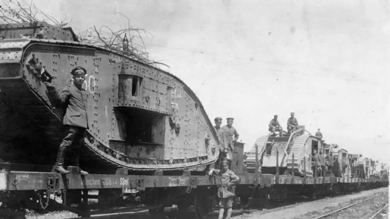 Een zwart-witfoto van een oude tank op een treinwagon, met soldaten erop en ernaast staand.