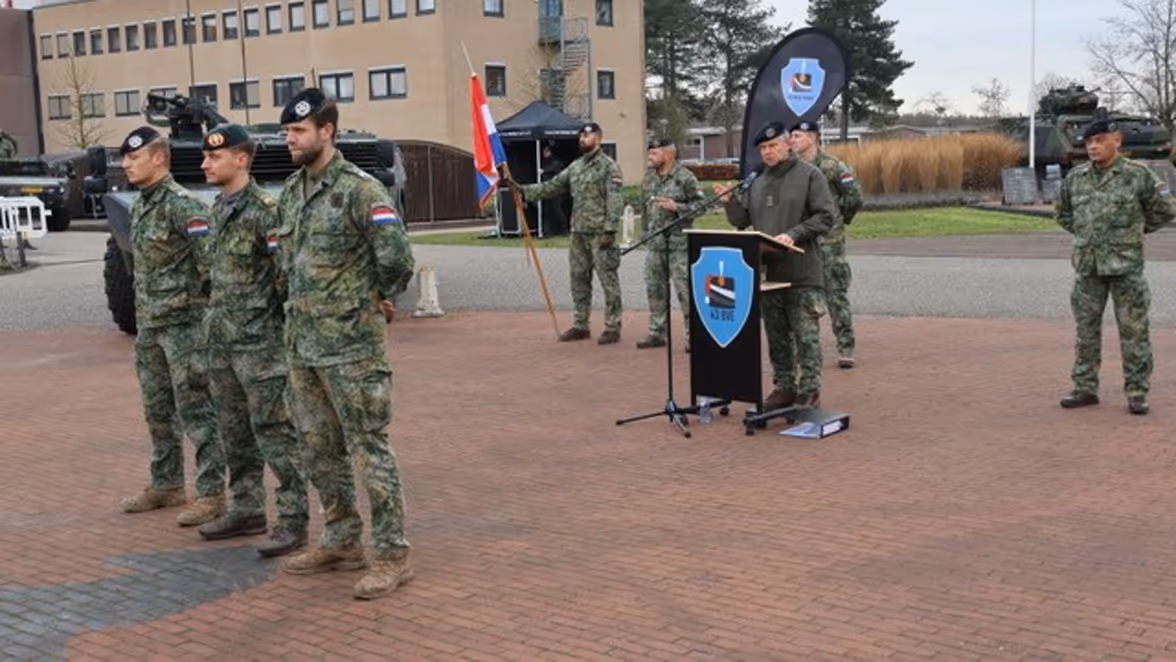 Militairen in uniform tijdens een buitenplechtigheid. Een persoon spreekt achter een lessenaar, met een Nederlands en een eenheidsvaandel op de achtergrond.