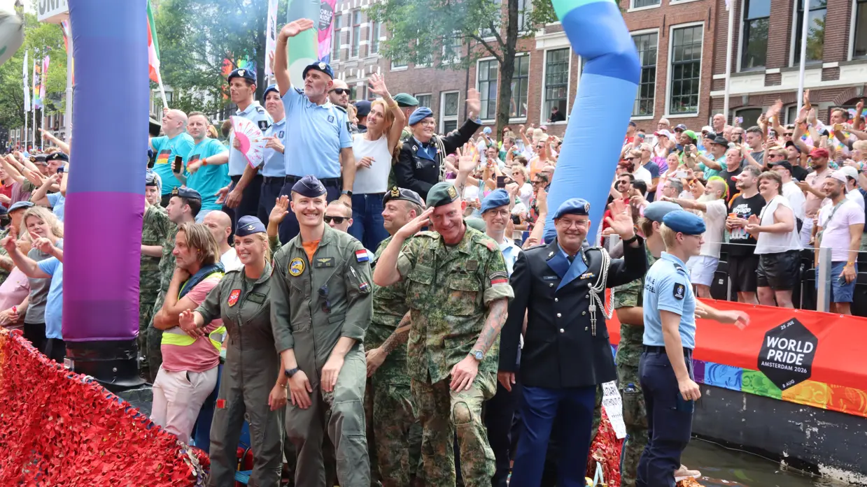 Een kleurrijke boot met mensen in uniformen en feestkleding vaart tijdens World Pride Amsterdam 2026, omringd door juichende toeschouwers.    