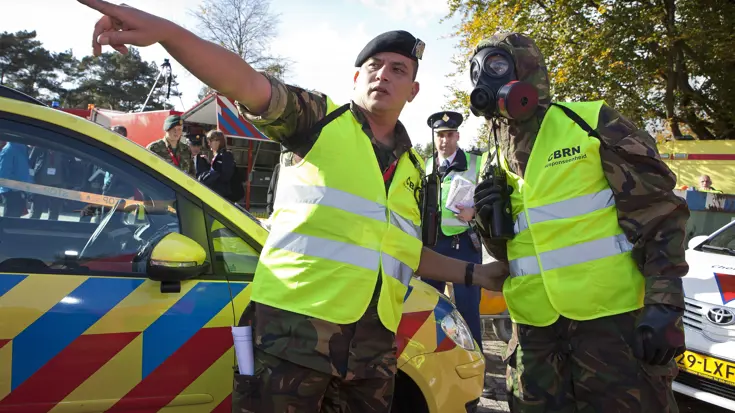 Een militair van de CBRN Respons Eenheid met beschermende uitrusting wordt naar een oefenramplocatie gewezen.