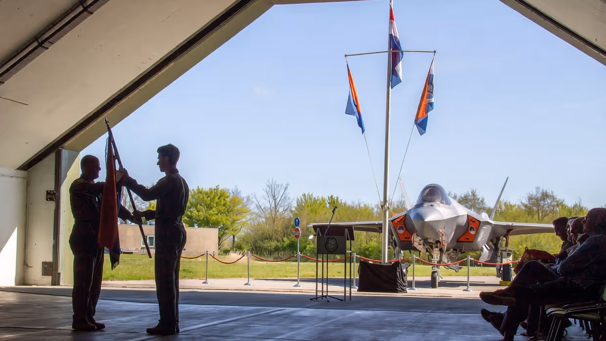 Scherders neemt de squadronvlag in ontvangst uit handen van Van de Graaf. Rechts van hen zit publiek. Op de achtergrond, net buiten de geopende shelter, staat een F-35 en een mast met drie vlaggen.