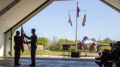 Scherders neemt de squadronvlag in ontvangst uit handen van Van de Graaf. Rechts van hen zit publiek. Op de achtergrond, net buiten de geopende shelter, staat een F-35 en een mast met drie vlaggen.