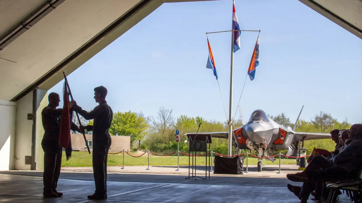 Scherders neemt de squadronvlag in ontvangst uit handen van Van de Graaf. Rechts van hen zit publiek. Op de achtergrond, net buiten de geopende shelter, staat een F-35 en een mast met drie vlaggen.