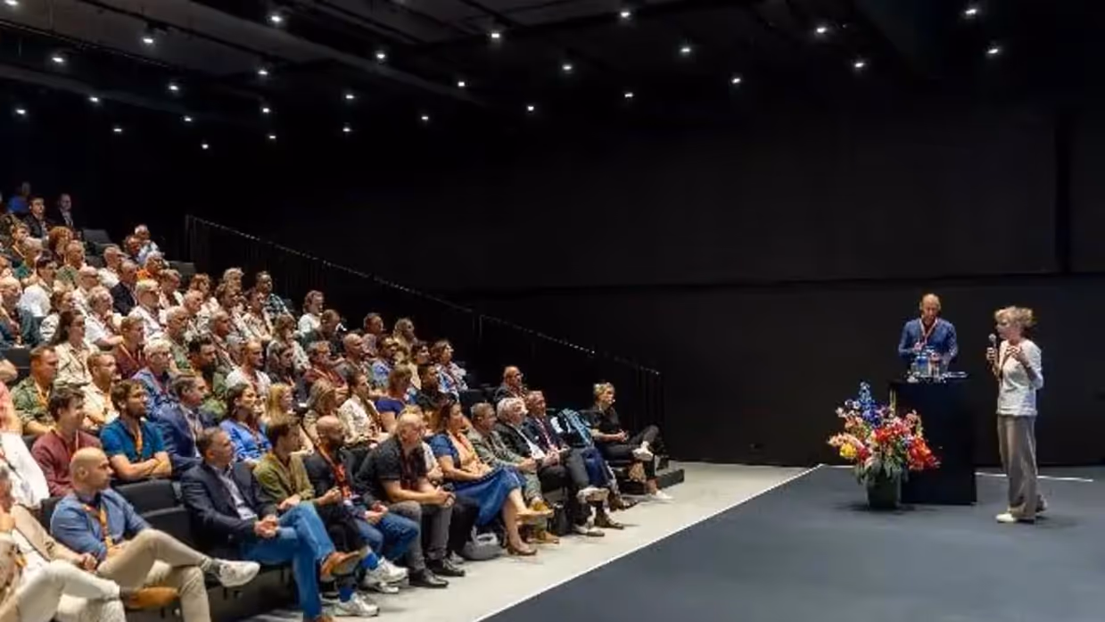  Een gevulde zaal. De aanwezigen kijken naar een podium, waarop een man en vrouw met microfoon staan.