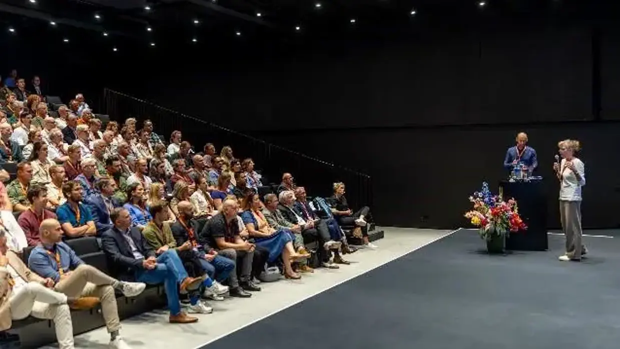  Een gevulde zaal. De aanwezigen kijken naar een podium, waarop een man en vrouw met microfoon staan.