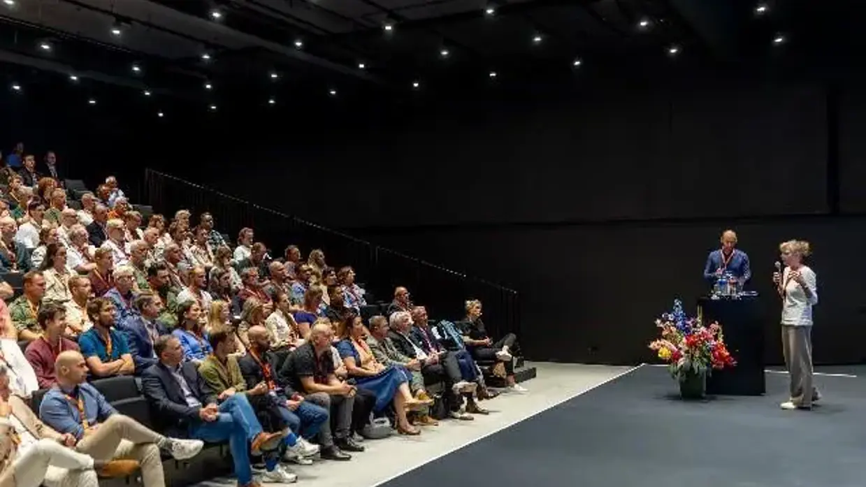  Een gevulde zaal. De aanwezigen kijken naar een podium, waarop een man en vrouw met microfoon staan.