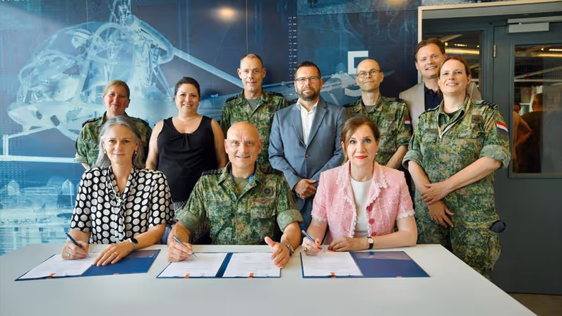 Generaal Steur zittend te midden van twee dames in burgerkleding. Alle drie hebben een pen in de hand en ondertekeningspapieren voor zich op tafel. Achter hen staan vier militairen in gevechtstenue en drie mensen in burgerkleding.