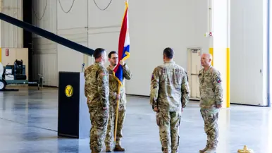 Een militair staat met een vlag in de hand tegenover een andere militair in een hangaar. Aan weerszijden staat een militair, de ene is Nederlands, de andere Amerikaans. Alleen dragen het gevechtstenue.