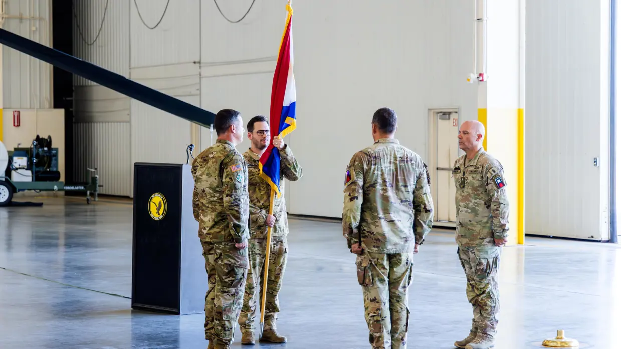 Een militair staat met een vlag in de hand tegenover een andere militair in een hangaar. Aan weerszijden staat een militair, de ene is Nederlands, de andere Amerikaans. Alleen dragen het gevechtstenue.