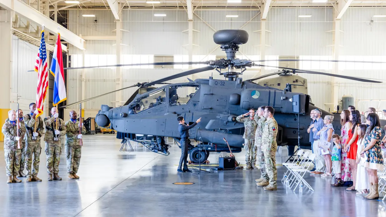 Ceremoniële bijeenkomst in een hangaar tegen de achtergrond van een Apache. Links staan vijf militairen onder wie er twee een lang wapen dragen en twee de Nederlandse en Amerikaanse vlag. Rechts staan vier militairen in de houding, achter hen staand publiek voor stoelen met de rechterhand op de linkerborst. In het midden zingt een tiener door een microfoon.