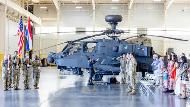 Ceremoniële bijeenkomst in een hangaar tegen de achtergrond van een Apache. Links staan vijf militairen onder wie er twee een lang wapen dragen en twee de Nederlandse en Amerikaanse vlag. Rechts staan vier militairen in de houding, achter hen staand publiek voor stoelen met de rechterhand op de linkerborst. In het midden zingt een tiener door een microfoon.