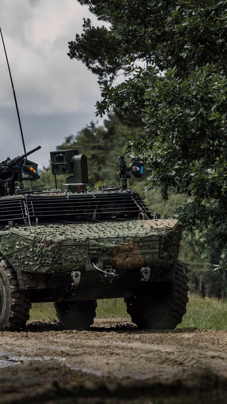 Een Fennek-voertuig met een camouflagenet rijdt door een beboste omgeving.