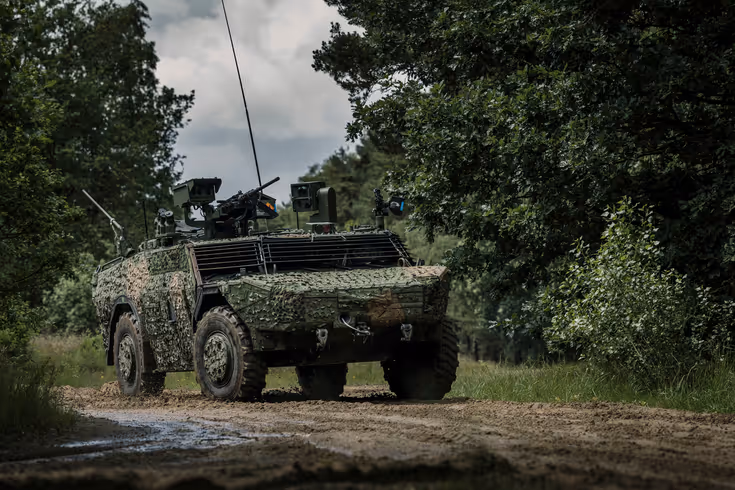 Een Fennek-voertuig met een camouflagenet rijdt door een beboste omgeving.