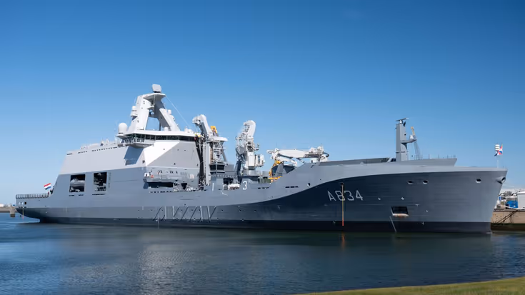 Een grijs marineschip ligt aan de kade. Op de voorgrond een grasstrook.