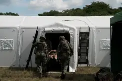 2 militairen op de rug gezien dragen een brancard met een gewonde een witte tent in.