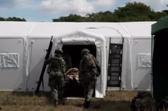 2 militairen op de rug gezien dragen een brancard met een gewonde een witte tent in.