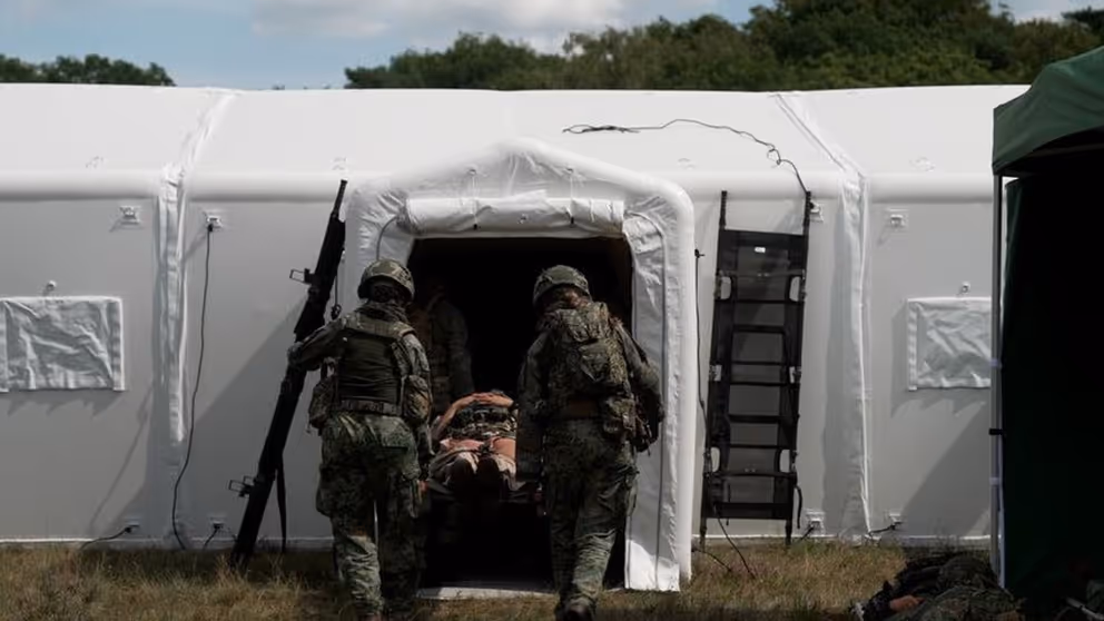 2 militairen op de rug gezien dragen een brancard met een gewonde een witte tent in.