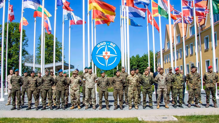 Een groep militairen staat, met de handen op de rug, opgesteld. Boven hen wapperen vlaggen van verschillende landen.