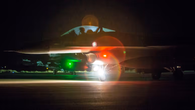 Nachtfoto van een taxiënde F-16 waarbij een groene en rode lamp fel in de camera schijnt.
