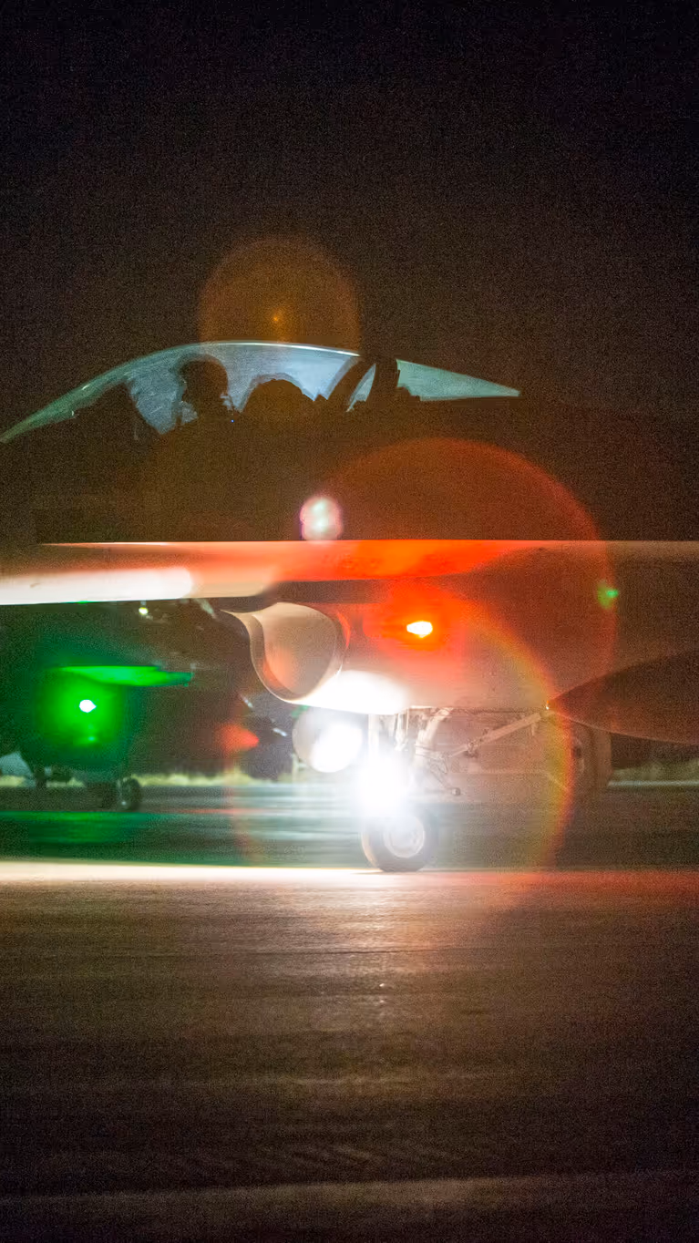 Nachtfoto van een taxiënde F-16 waarbij een groene en rode lamp fel in de camera schijnt.