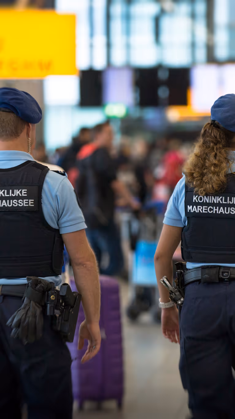 Twee marechaussees tussen het publiek op Schipol, op de rug gezien.