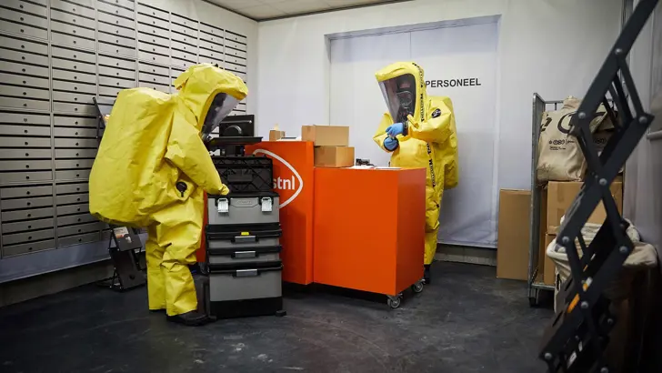 Personen in beschermende gele pakken onderzoeken een postkantoor.