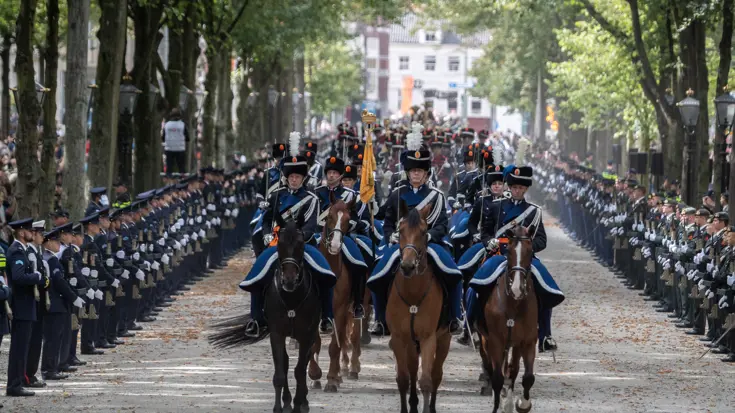 Marechaussees in ceremonieel tenue op paarden. Links en rechts langs de weg een ere-afzetting.