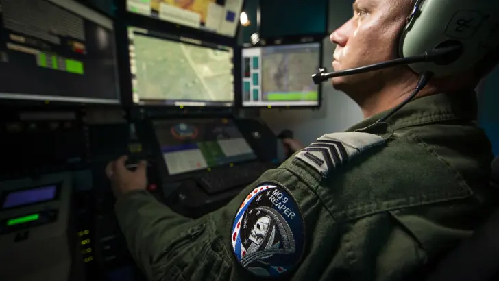 Een vlieger en sensoroperator vliegen een toestel vanachter beeldschermen. Op de mouw een MQ-9 Reaper-embleem met Magere Hein en de Friese vlag.