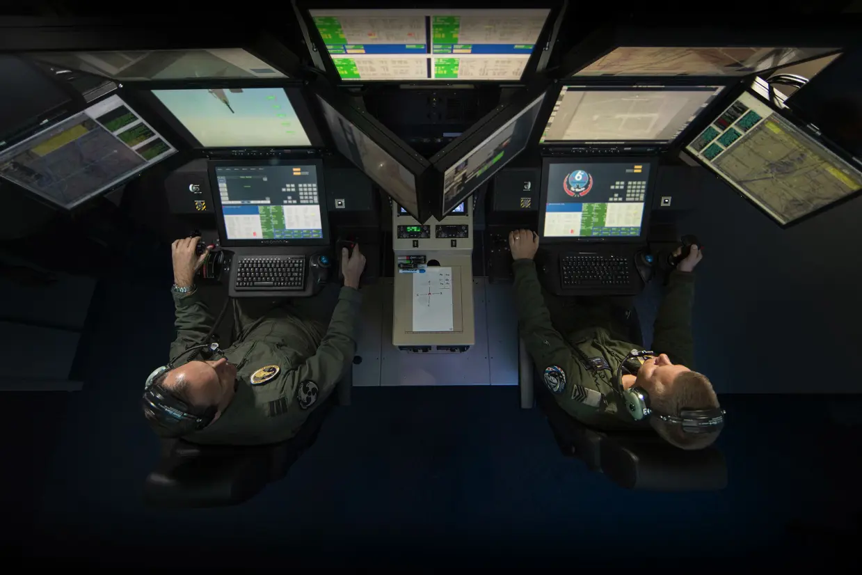 Een vlieger en sensoroperator zitten zij aan zij (van boven bezien) terwijl ze een toestel besturen vanachter beeldschermen.