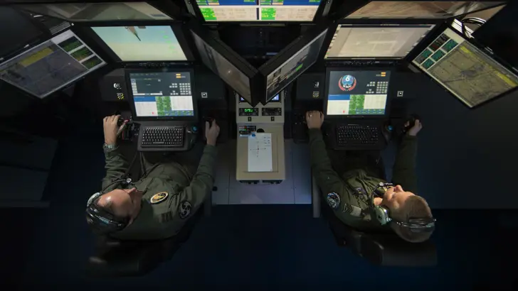 Een vlieger en sensoroperator zitten zij aan zij (van boven bezien) terwijl ze een toestel besturen vanachter beeldschermen.