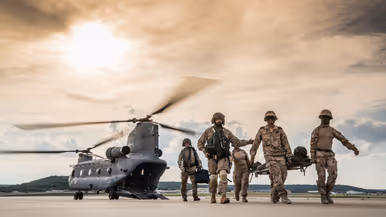 Militairen lopen over een platform met een gewonde collega op een brancard. Op de achtergrond staat een Chinook met draaiende rotorbladen en een geopende laadklep.