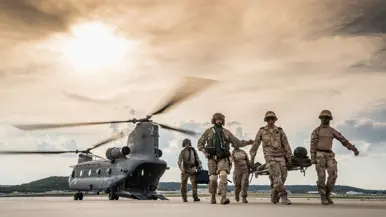 Militairen lopen over een platform met een gewonde collega op een brancard. Op de achtergrond staat een Chinook met draaiende rotorbladen en een geopende laadklep.