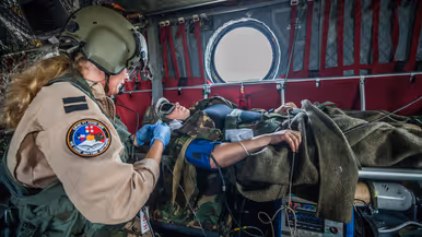 Een militair met blauwe handschoenen verzorgt een collega op een brancard in een helikopter. Op haar arm een patch met de woorden ‘ORAC Ft Hood 2019 – we care anytime anywere’.